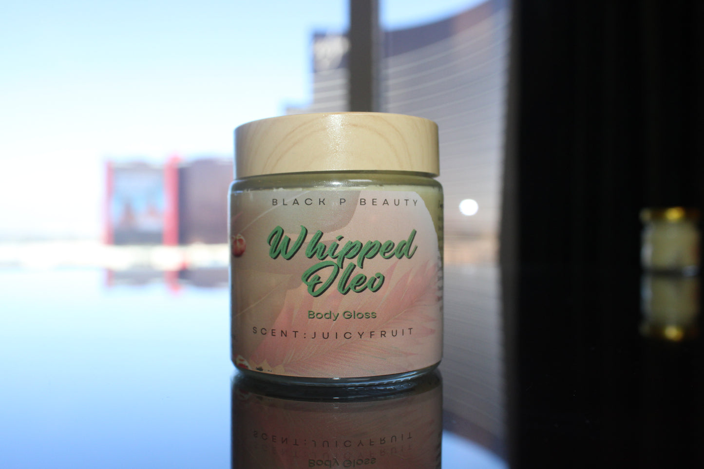 Whipped Oleo body gloss in a glass jar with wood effect top sitting on a luxury reflective table. Body butter body glaze