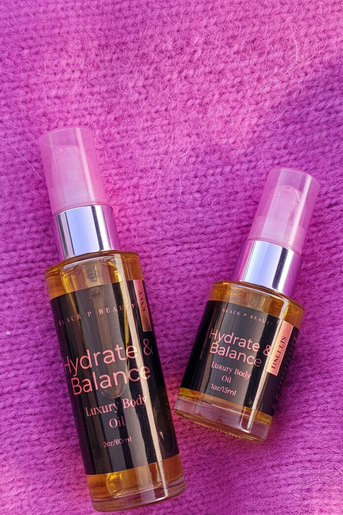 Two clear spray bottles of Black Op Body's Hydrate & Balance Luxury Body Oil with pink caps and black labels, placed on a textured hot pink background.