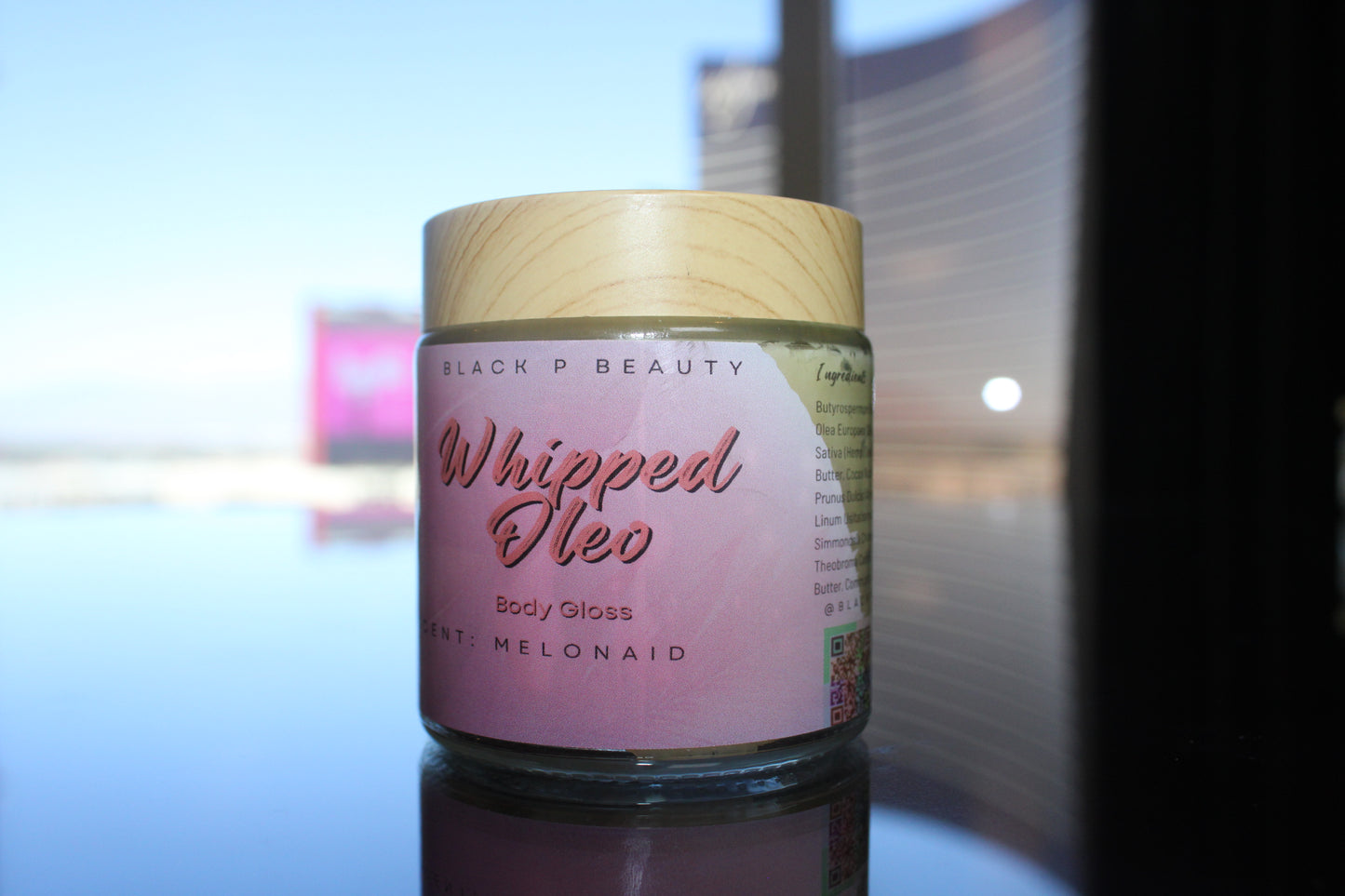 Whipped Oleo body gloss in a glass jar with wood effect top sitting on a luxury reflective table. Body butter body glaze